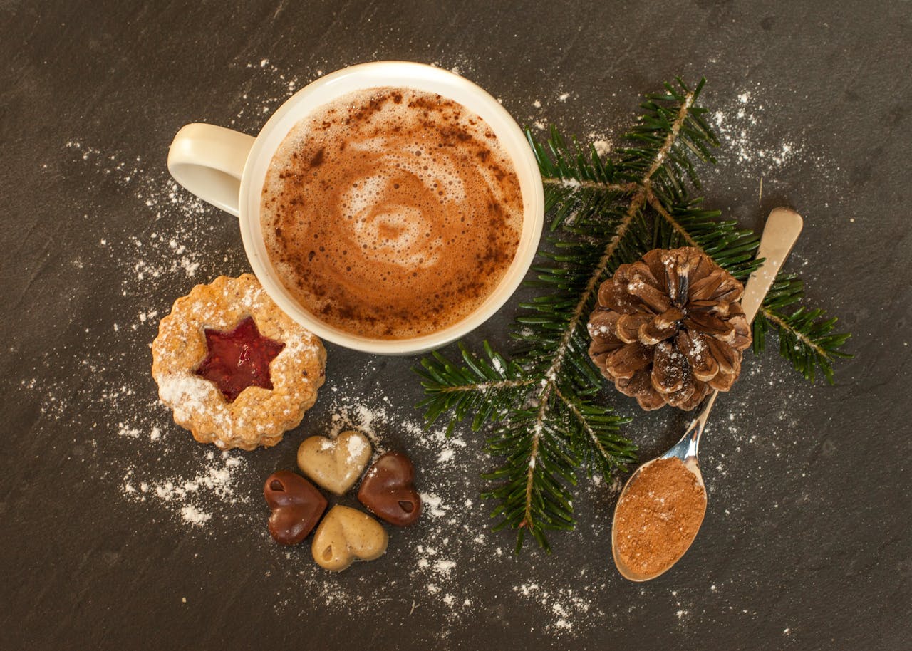White mug of hot chocolate with festive cookies, pinecone, and cinnamon on a dark surface.