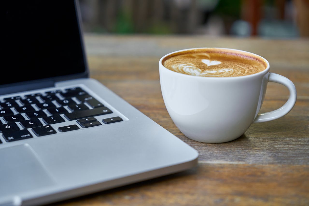 About Close-up of a latte coffee with heart art next to a laptop on a rustic wooden table.